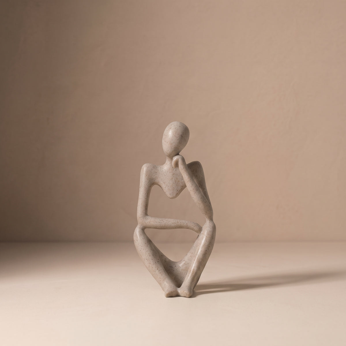 Seated Figure, Form I, in matte speckled stone. Abstract seated sculpture, upright with one arm crossing the body, on a pale plaster floor.