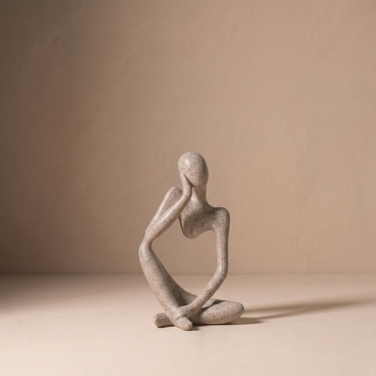 Seated Figure, Form II, in matte speckled stone. Abstract seated sculpture resting its chin on one hand, on a pale plaster floor.