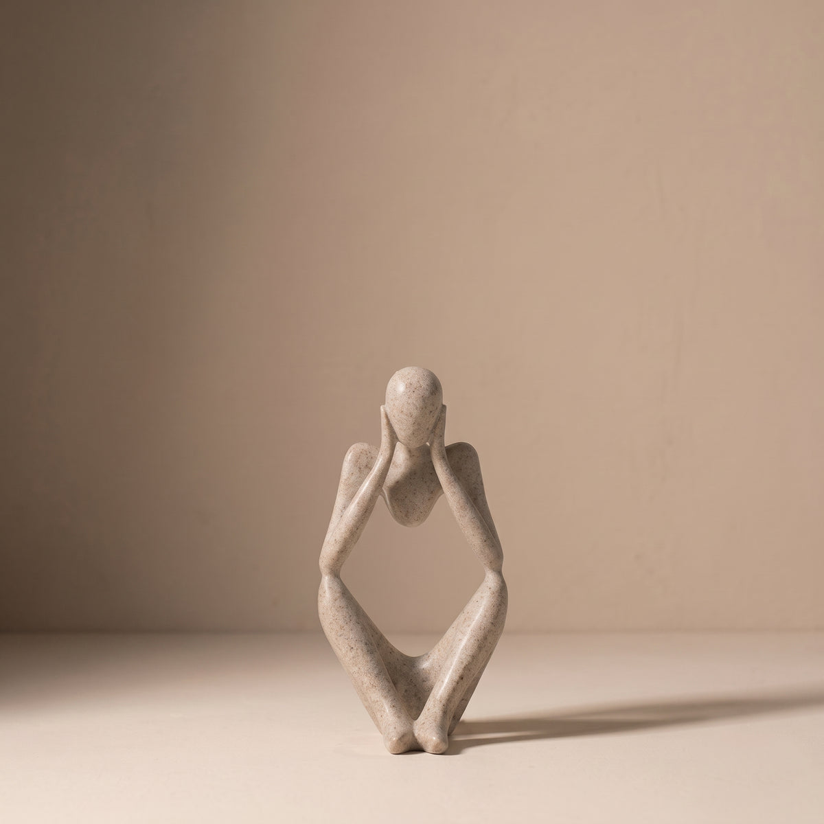 Seated Figure, Form III, in matte speckled stone. Abstract seated sculpture with both hands lifted to the face, on a pale plaster floor.
