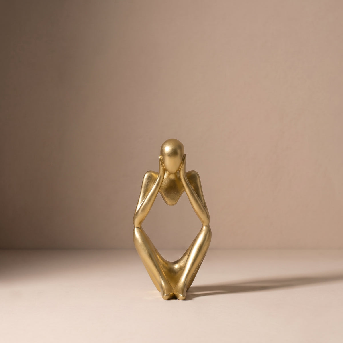 Seated Figure, Form III, in brushed satin gold. Abstract seated sculpture with both hands lifted to the face, on a pale plaster floor.