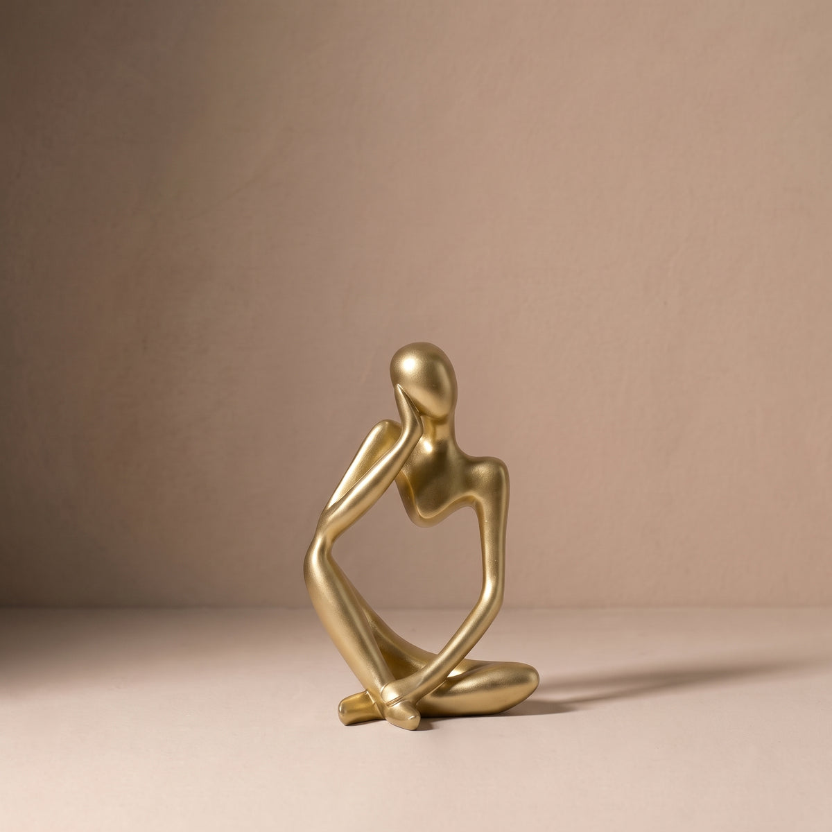 Seated Figure, Form II, in brushed satin gold. Abstract seated sculpture resting its chin on one hand, on a pale plaster floor.