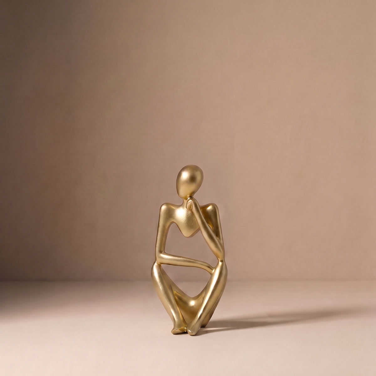 Seated Figure, Form I, in brushed satin gold. Abstract seated sculpture, upright with one arm crossing the body, on a pale plaster floor.